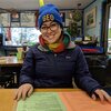Grad student Ashli Anda sits in a restaurant before COVID and smiles at the camera; she is wearing a GEO beanie and a scarf and jacket