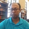 Alamgir Hossain sits in front of two bookshelves and windows during day time.